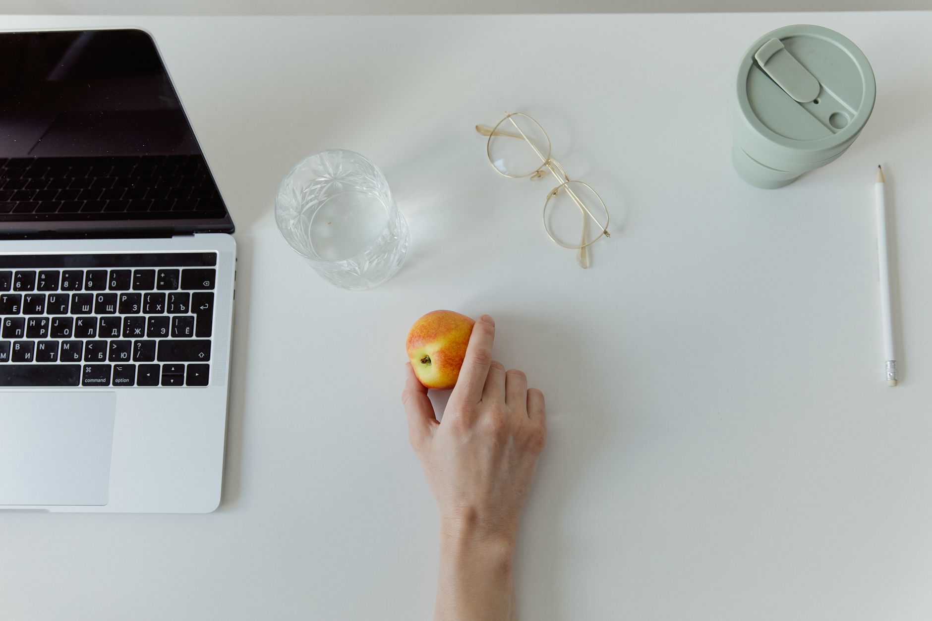 Overhead shot of a desk with a laptop open, a healthy snack (like an apple) beside it, and a hand reaching for the snack.